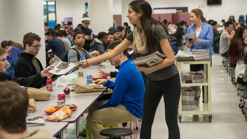 A girl hands out newspapers to students sitting at a lunch table in a school cafeteria. 