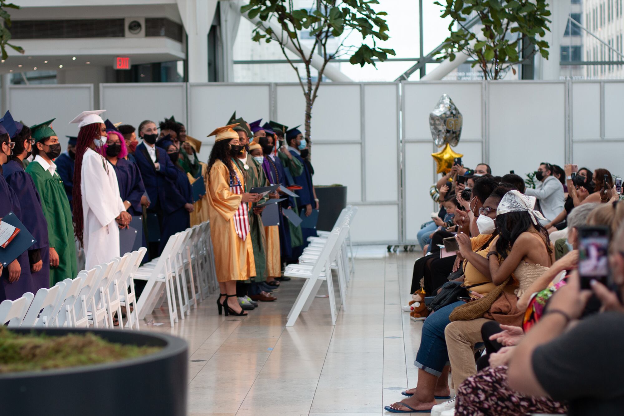 Graduating students stand and face their families that are sitting down as their families applaud in a large auditorium space.