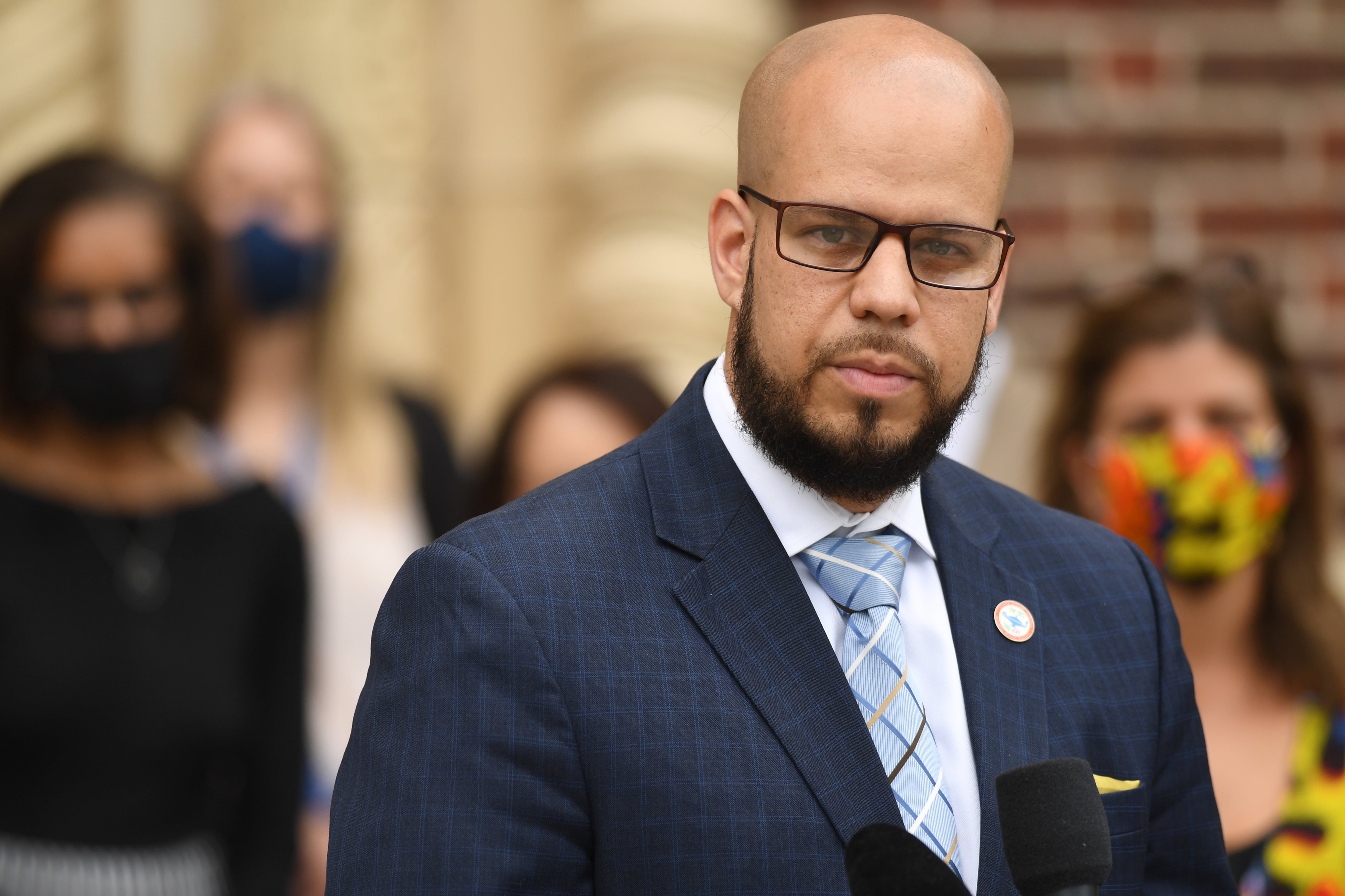 Dr. Alex Marrero, wearing a dark checkered suit jacket, white shirt, blue striped tie and a lapel pin.