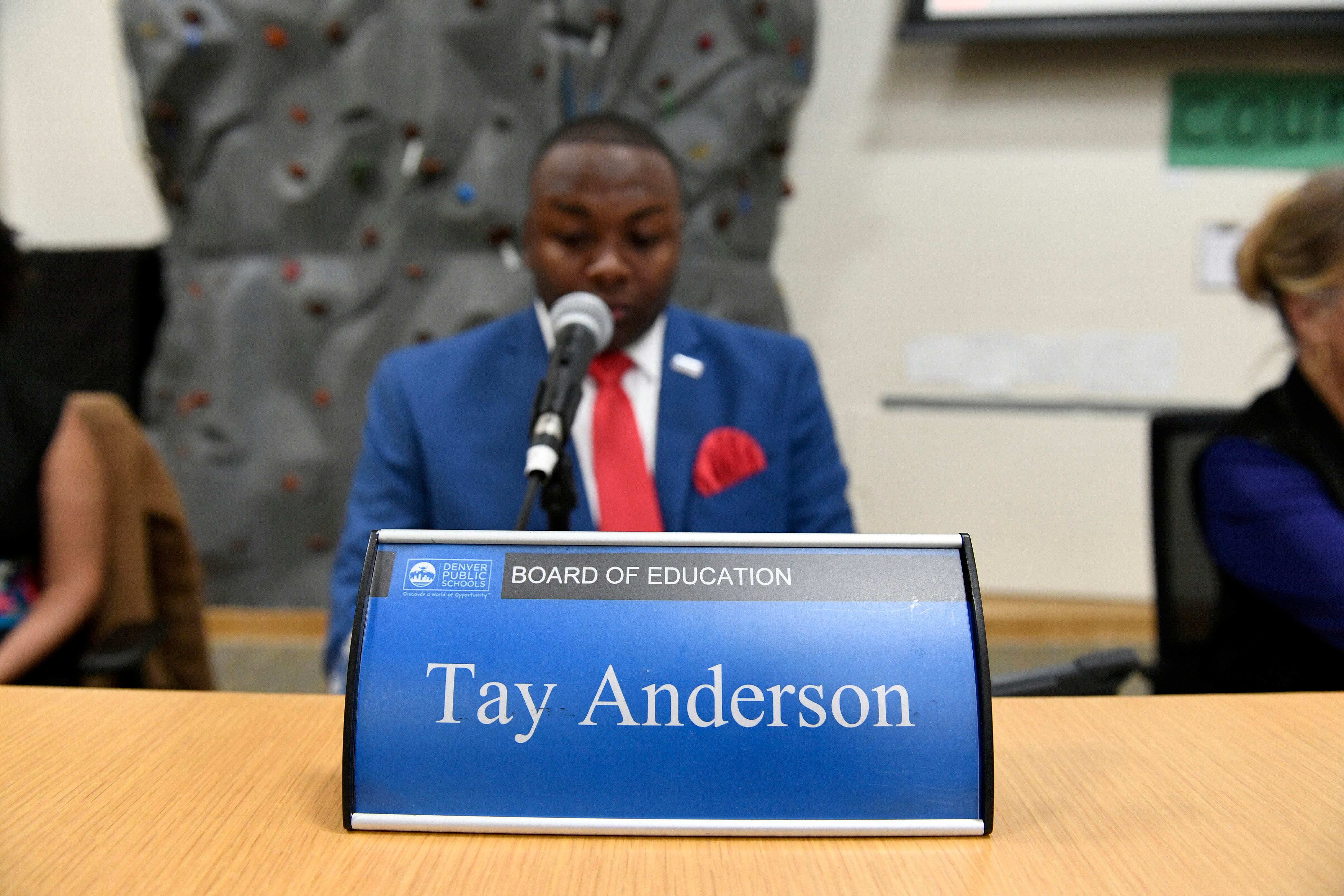 Tay Anderson, wearing a red tie and blue suit, sits behind a blue placard with his name typed in white letters.