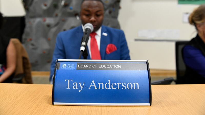 Tay Anderson, wearing a red tie and blue suit, sits behind a blue placard with his name typed in white letters.