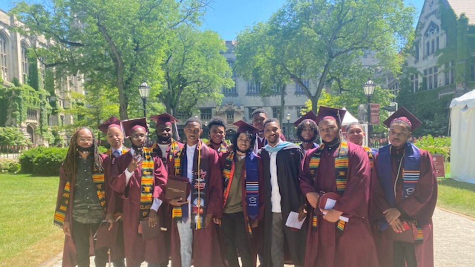 A group of high school graduates, all young Black men, wear maroon caps and gowns. They stand together with their mentor.
