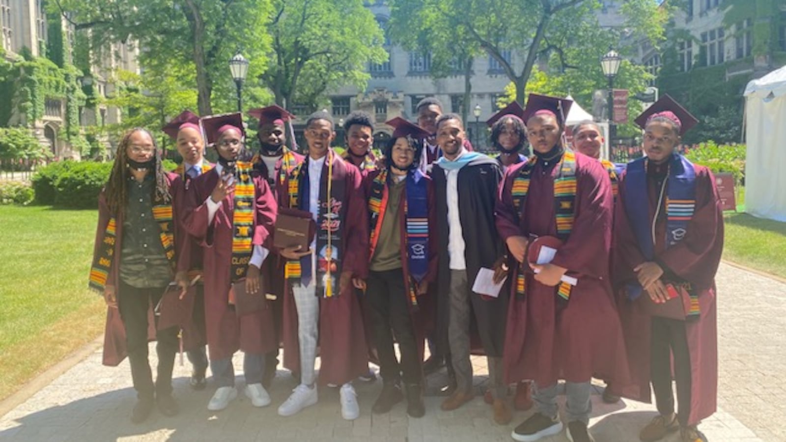 A group of high school graduates, all young Black men, wear maroon caps and gowns. They stand together with their mentor.