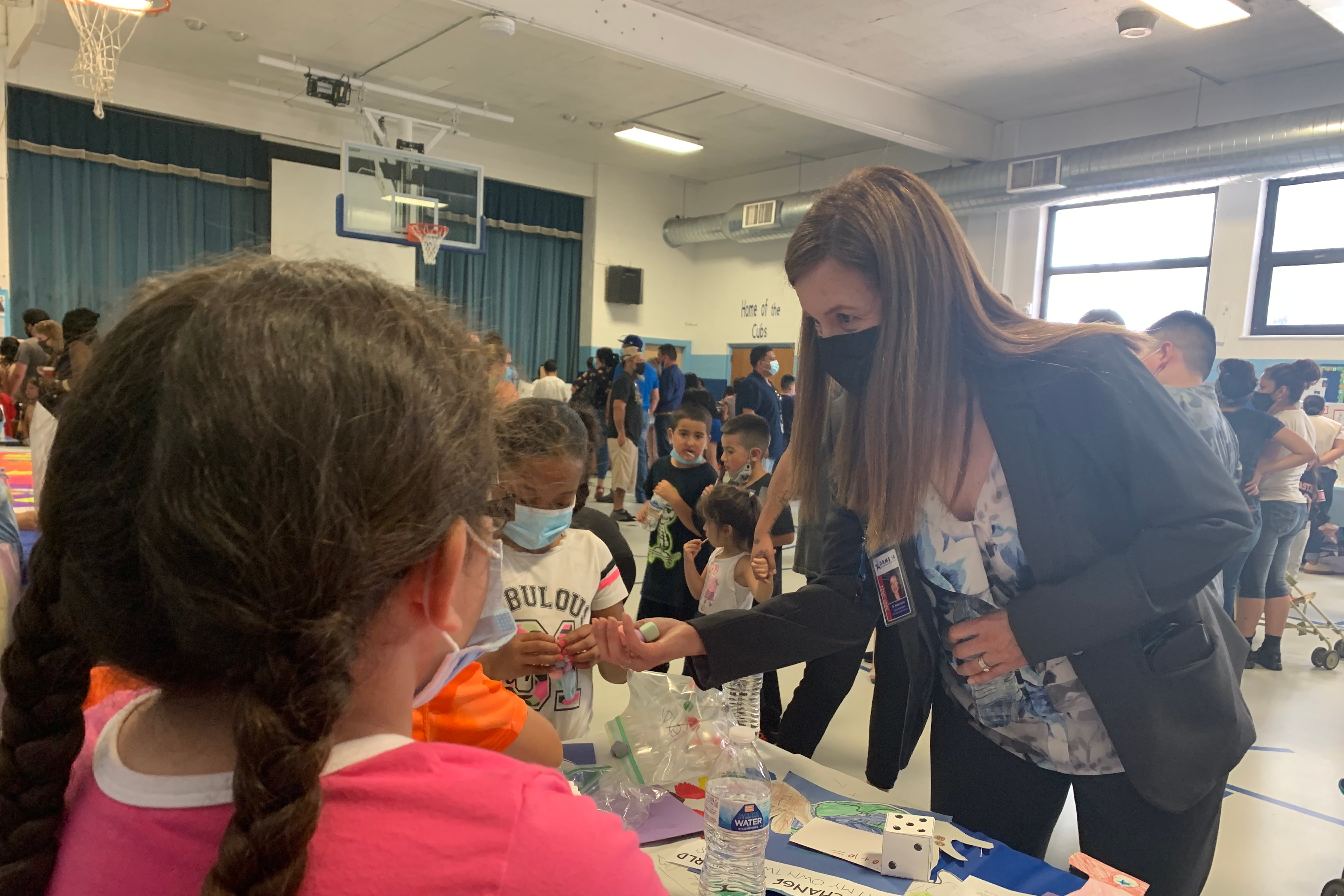 A masked woman in a suit leans in over a long table to talk with students in a crowded school gym.