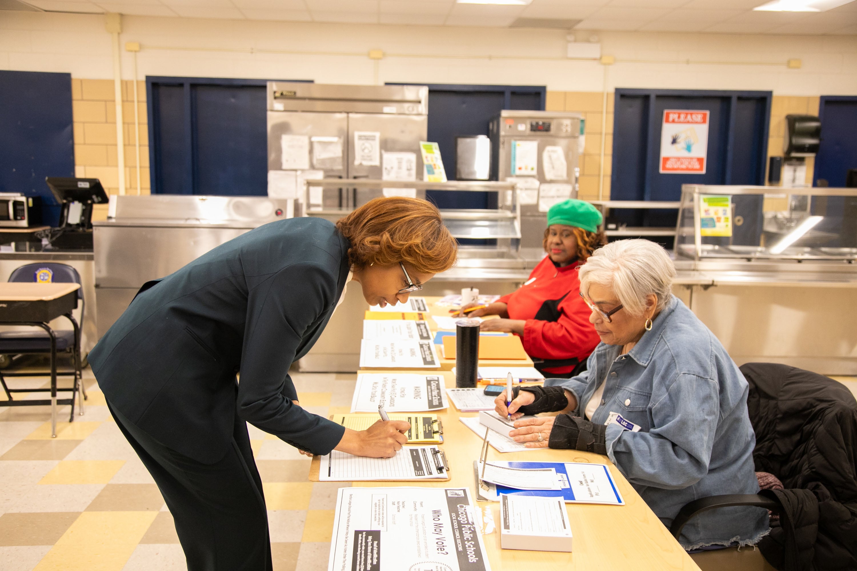 A photograph of a Black woman in a suit bending over a table filling out a piece of paper while two other women sit at the table and assist.