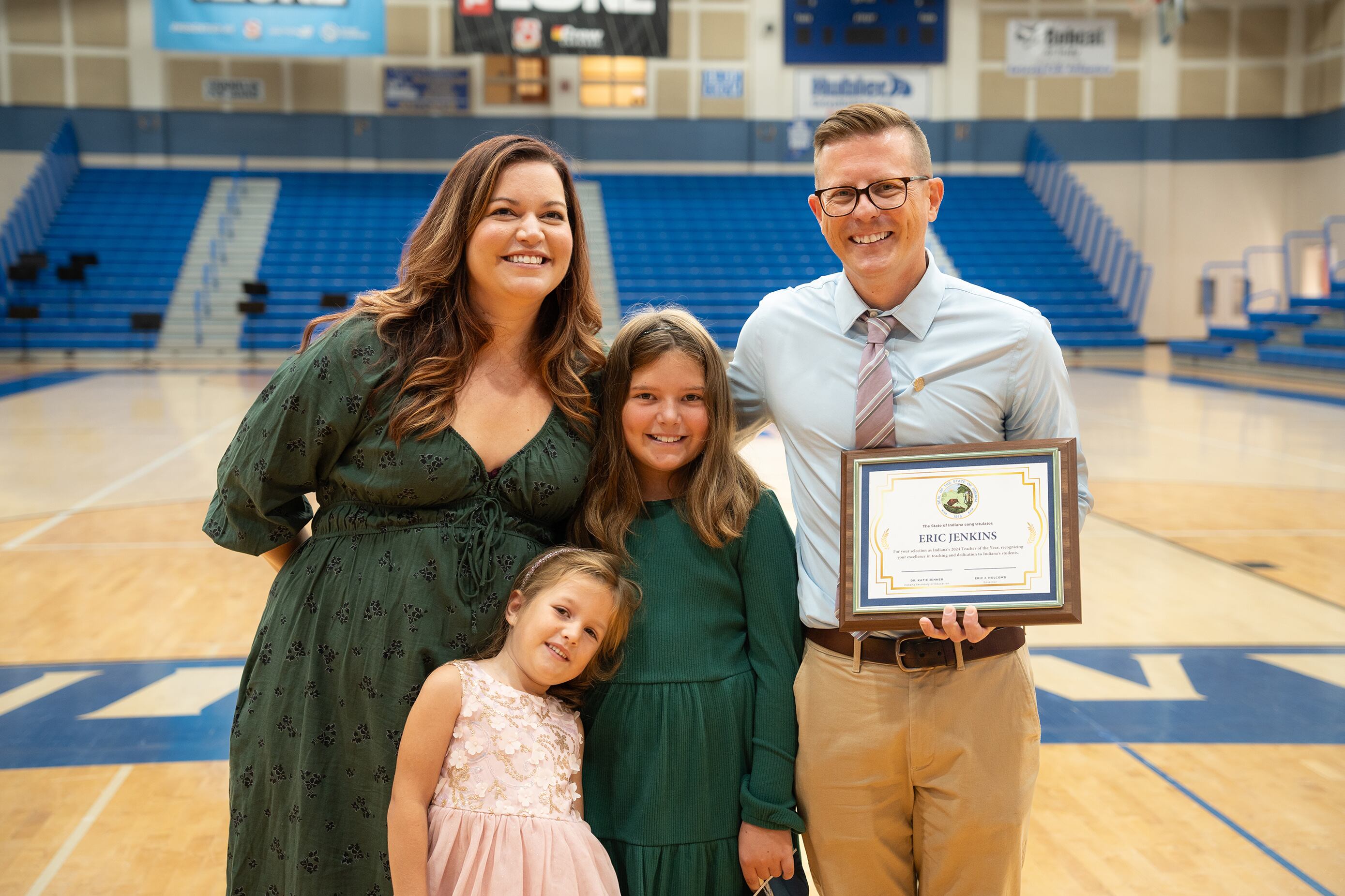Two adults and two children pose for a photograph while one adult holds a framed award. They are standing in a school gym.