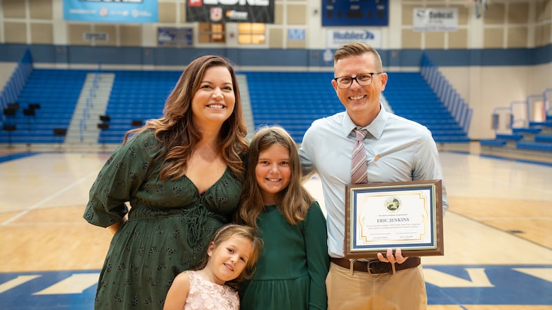 Two adults and two children pose for a photograph while one adult holds a framed award. They are standing in a school gym.