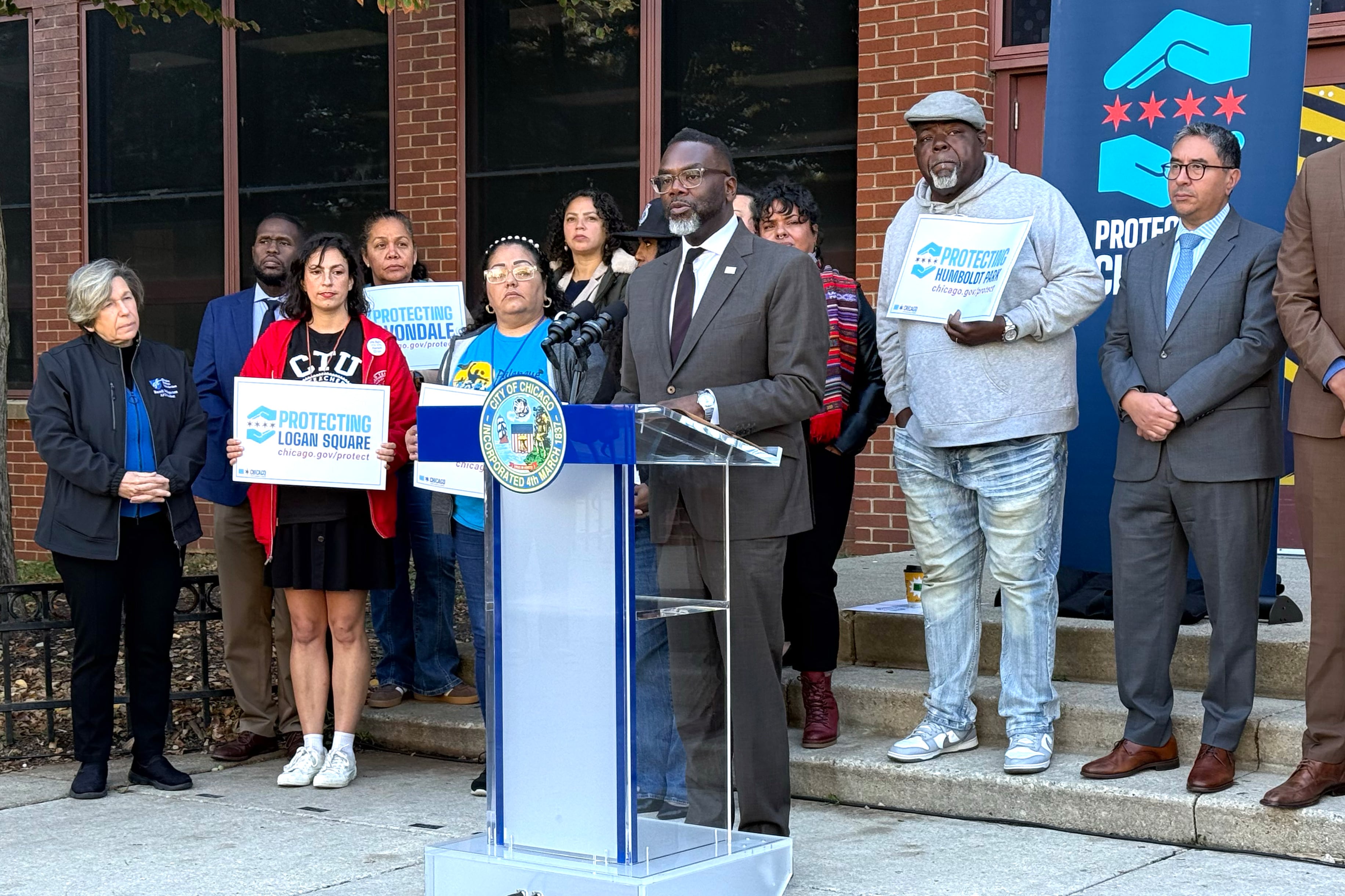 A man in a gray suit speaks while others, some holding signs, stand behind him.