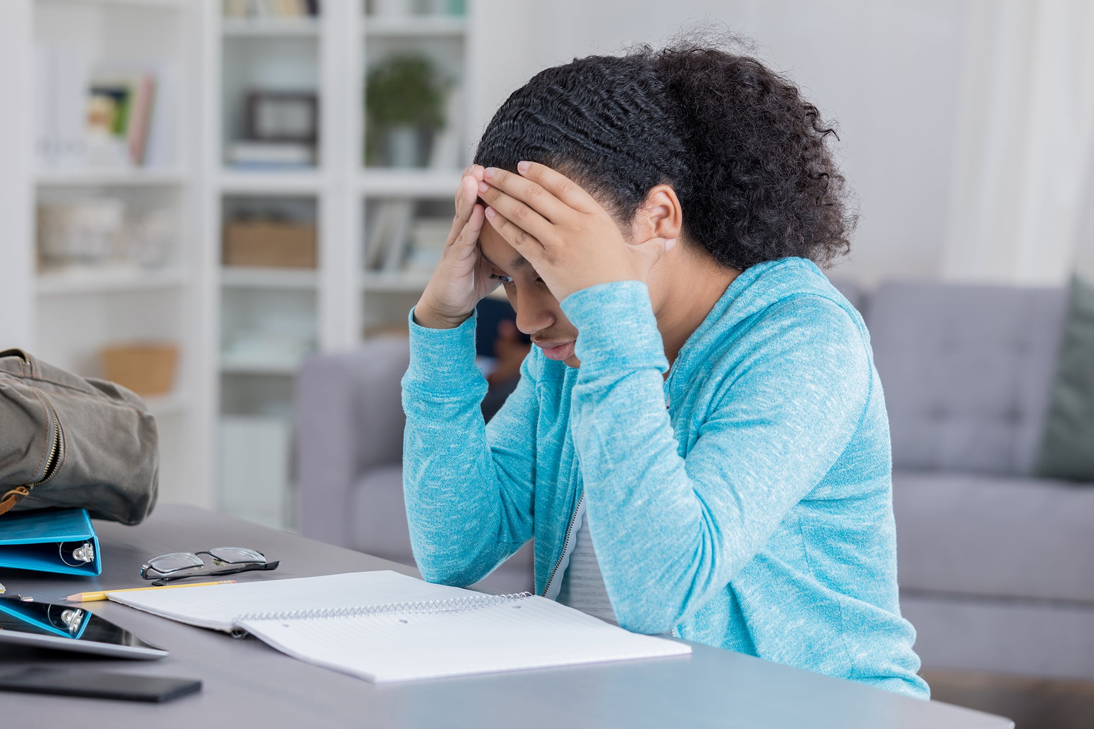 A high school girl with dark, curly hair and wearing a blue sweater sits at a table with her hands to her face in a stressed out way with a white notebook on the table in front of her.