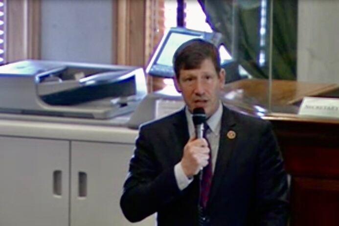 A man in a suit speaks into a microphone in front of a copy machine setting on top of a a cabinet.