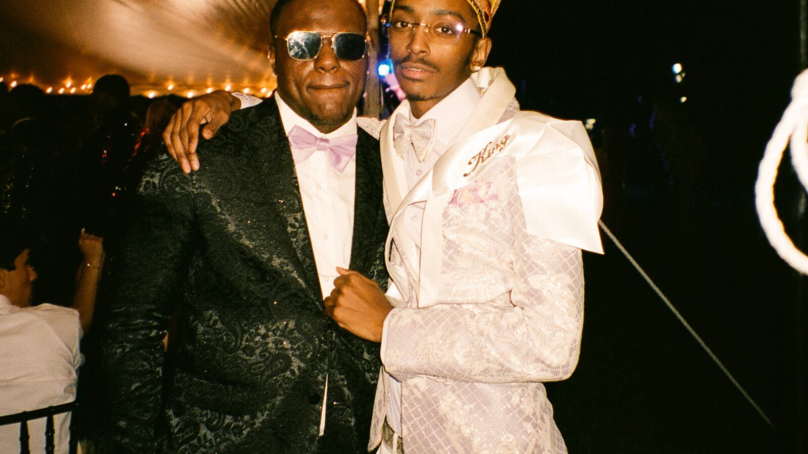 Two young men, one wearing a black tuxedo and another wearing a white tuxedo and gold crown, pose for a portrait together under a party tent at their prom.