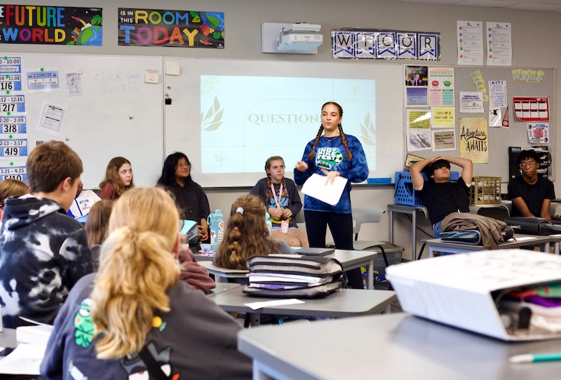 An eighth grade student stands up in the middle of a group of students sitting in chairs in a classroom.