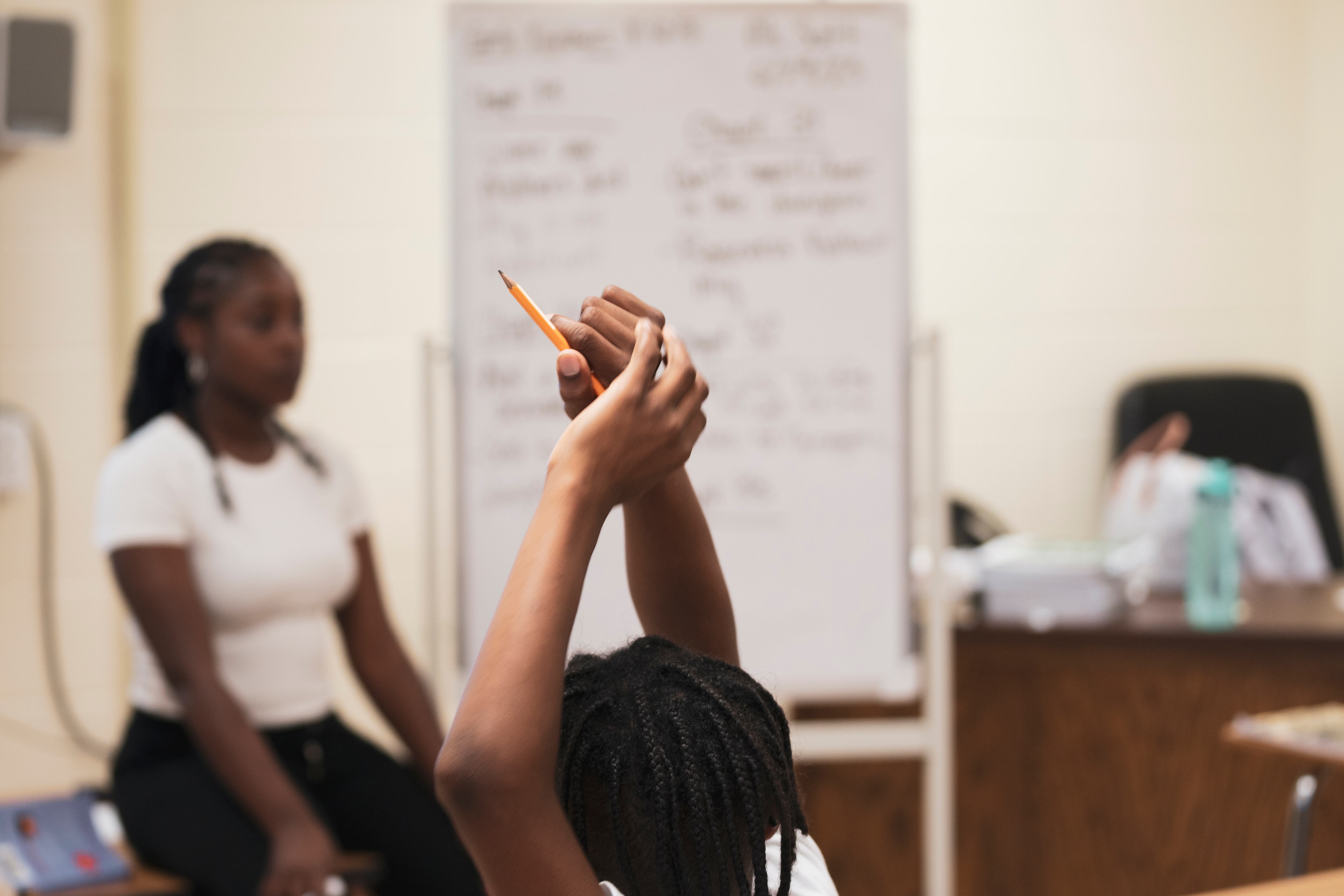 A close up of a student raising their hands with a teacher and a classroom in the background.
