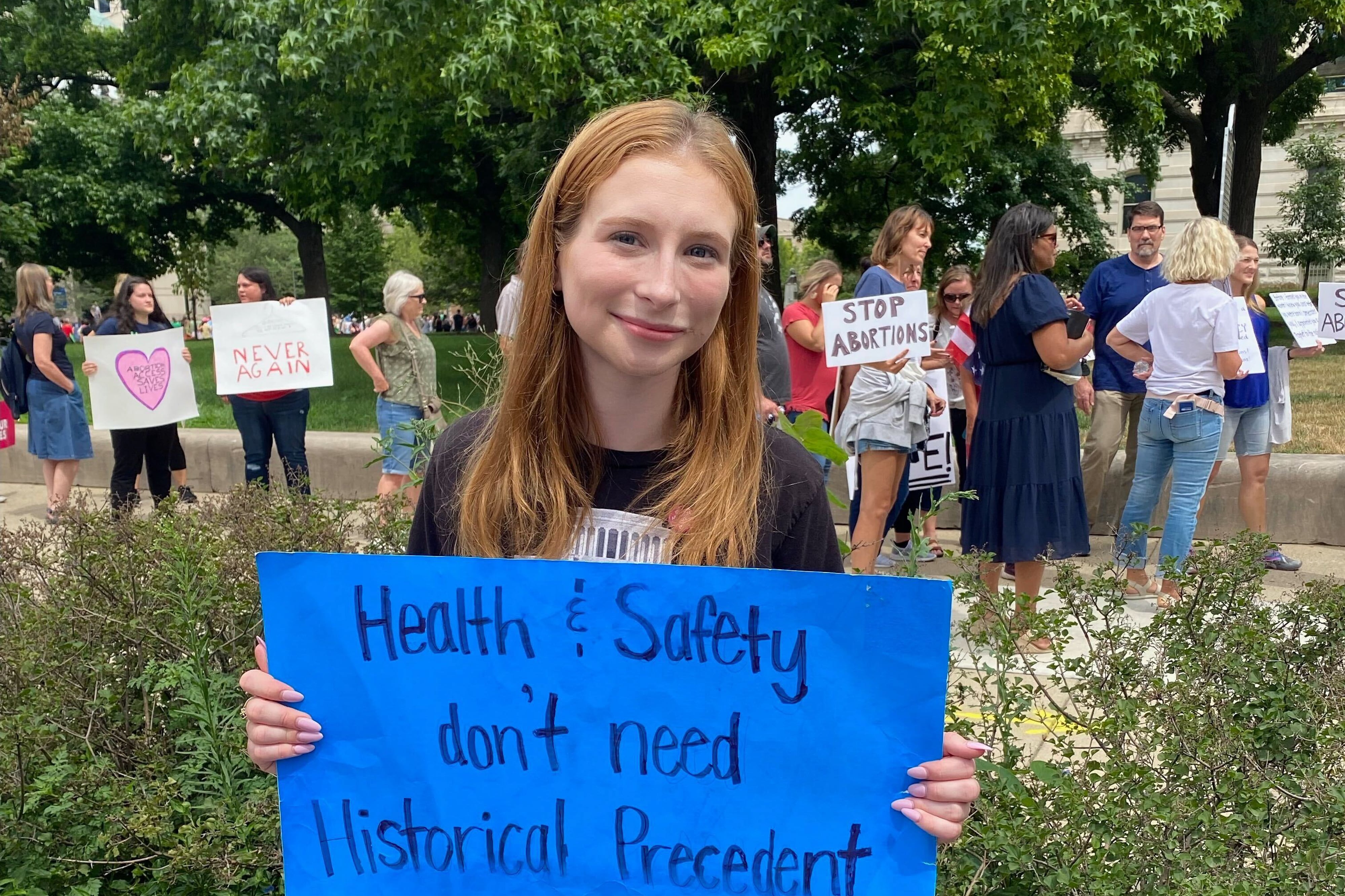 A young woman holds a blue sign and stands in front of a crowd of people holding signs. Her sign reads: “Health and safety don’t need historical precedent.”