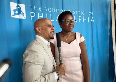 Two people in business clothes stand in front of a blue sheet background that reads " The School district of Philadelphia".