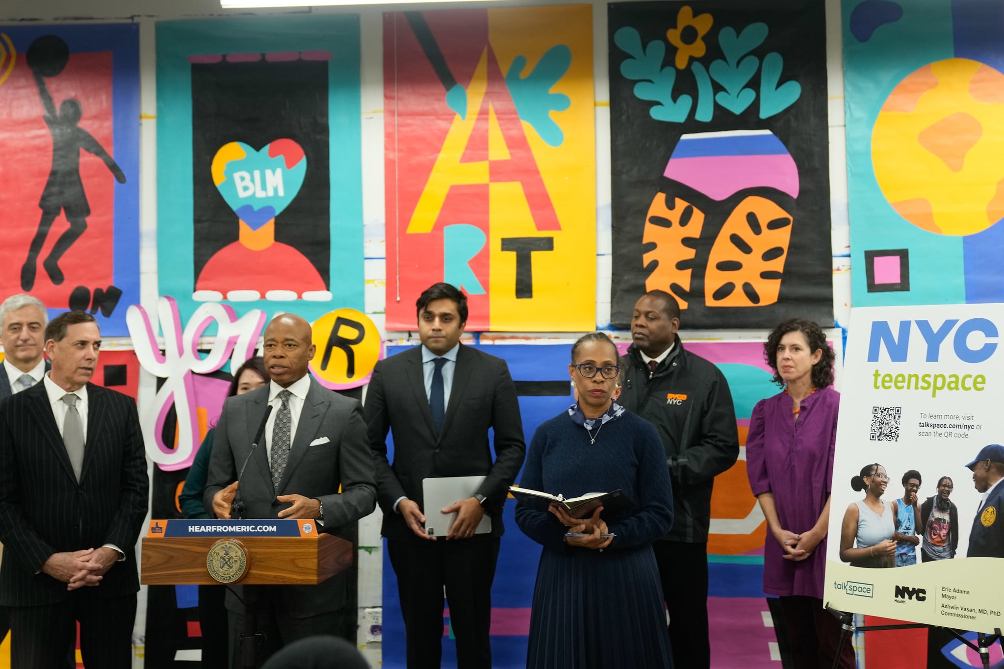 A group of men in suits and two women stand in front of a wall with bright mural on it. Mayor Adams is at a lectern.  A sign reads NYC TeenSpace.