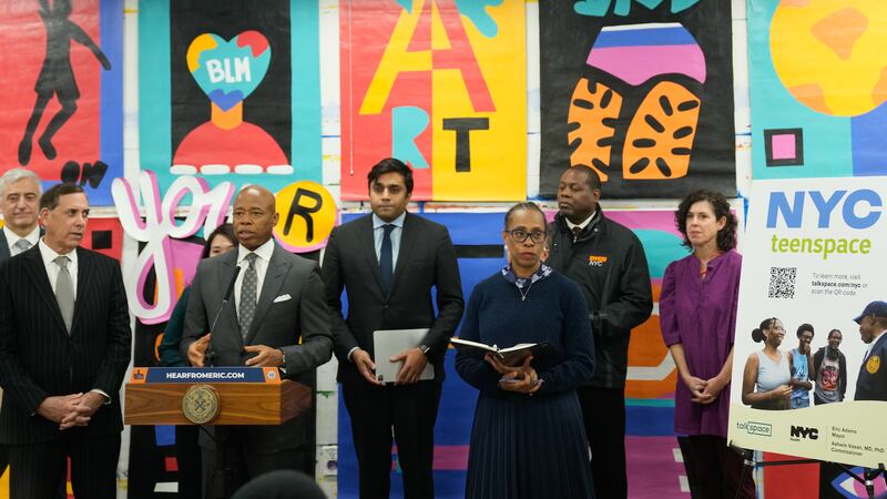 A group of men in suits and two women stand in front of a wall with bright mural on it. Mayor Adams is at a lectern.  A sign reads NYC TeenSpace.