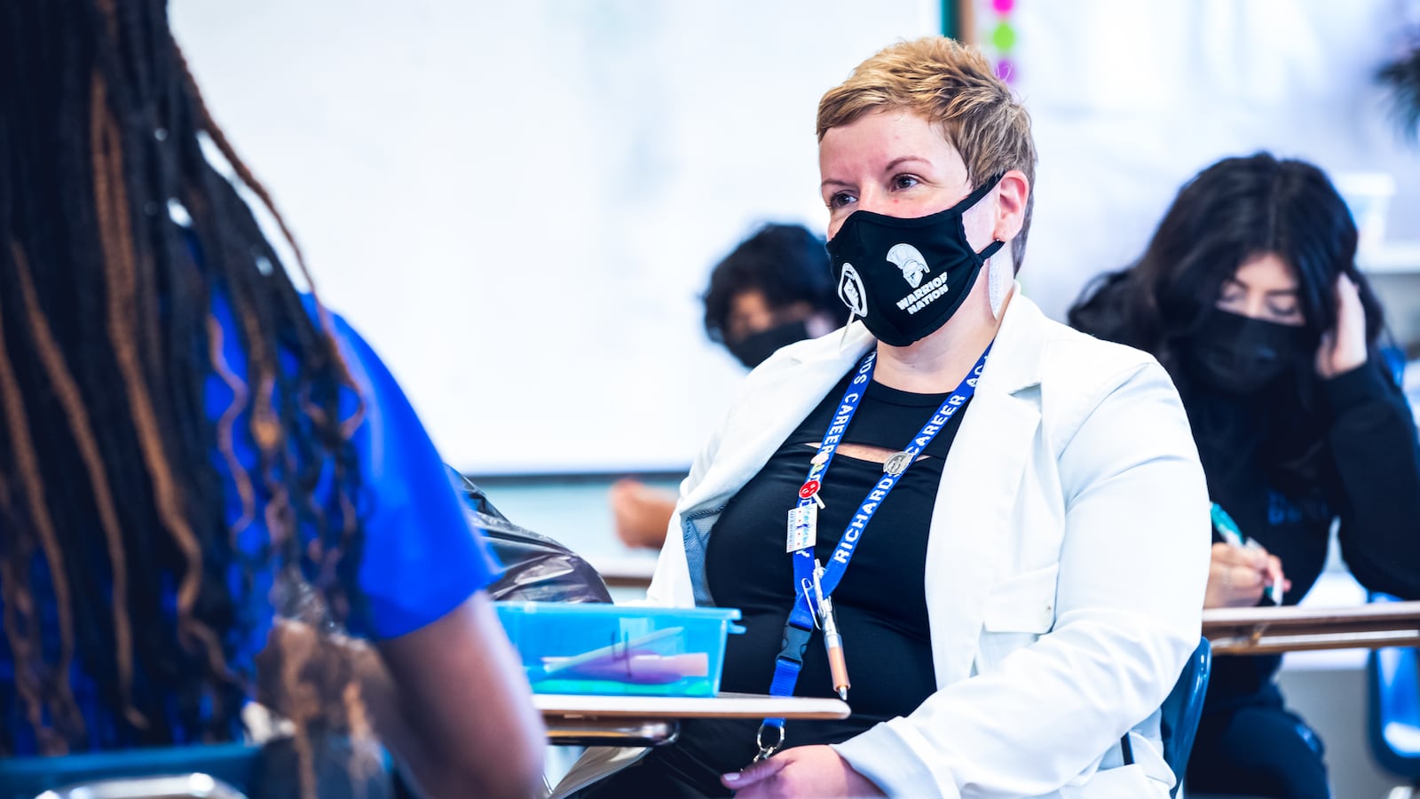 A woman in a face mask sits at a desk and speaks with a girl in a school classroom.