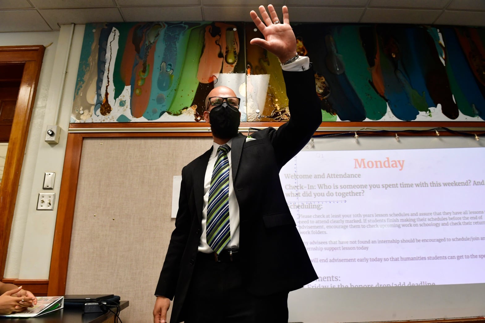 A man wearing a suit and tie and face mask stands in a classroom, waving at students.