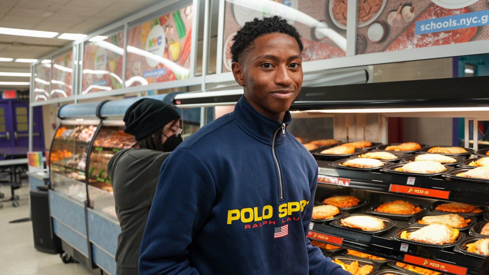 A high school student with short dark hair and wearing a blue sweater holds a tray of lunch food with another person looking at food in the background.
