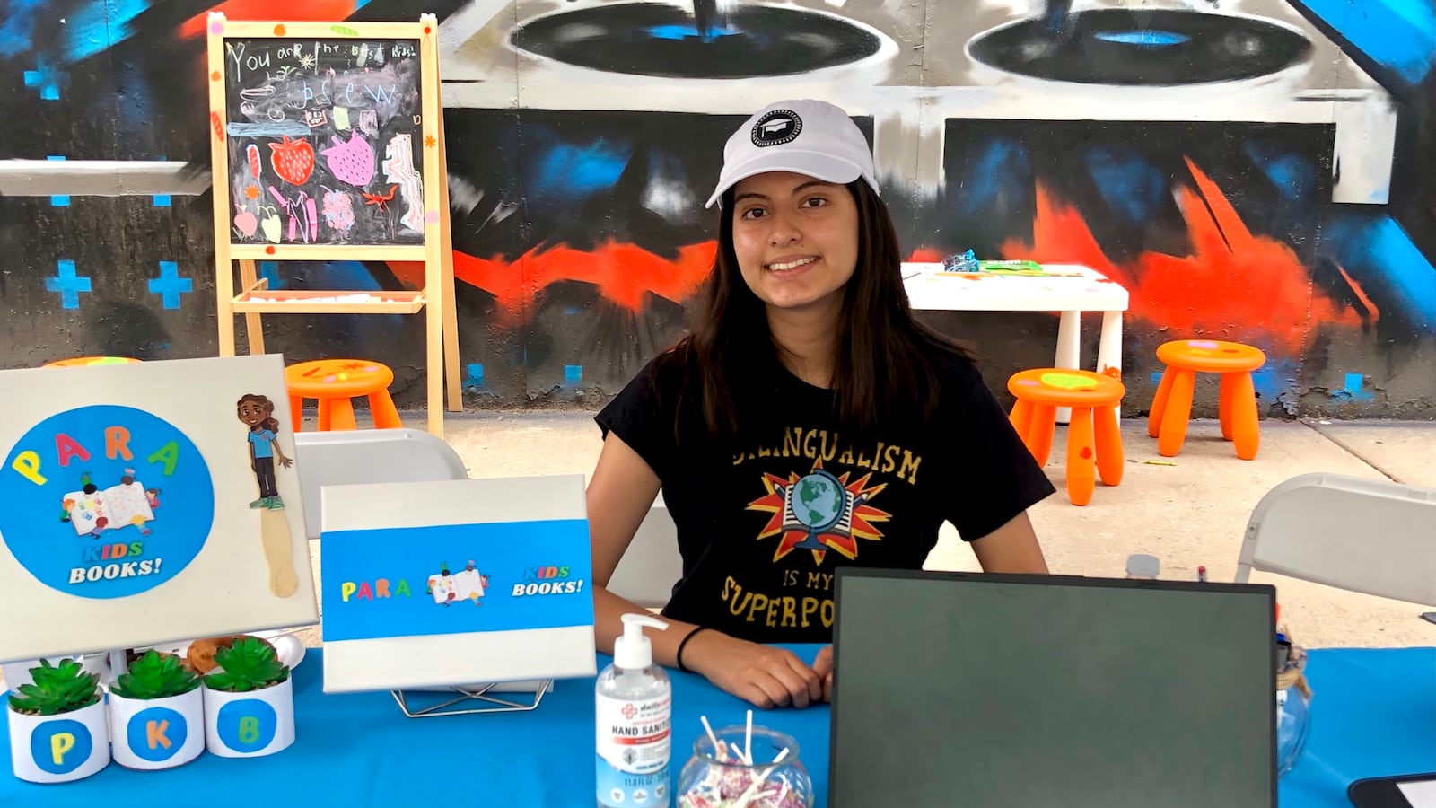 Young woman in a black t-shirt and white cap sits at a table surrounded by promotional materials.