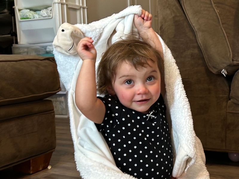 A 1-yr-old girl holds a blanket above her head while she sits on the floor of her home.