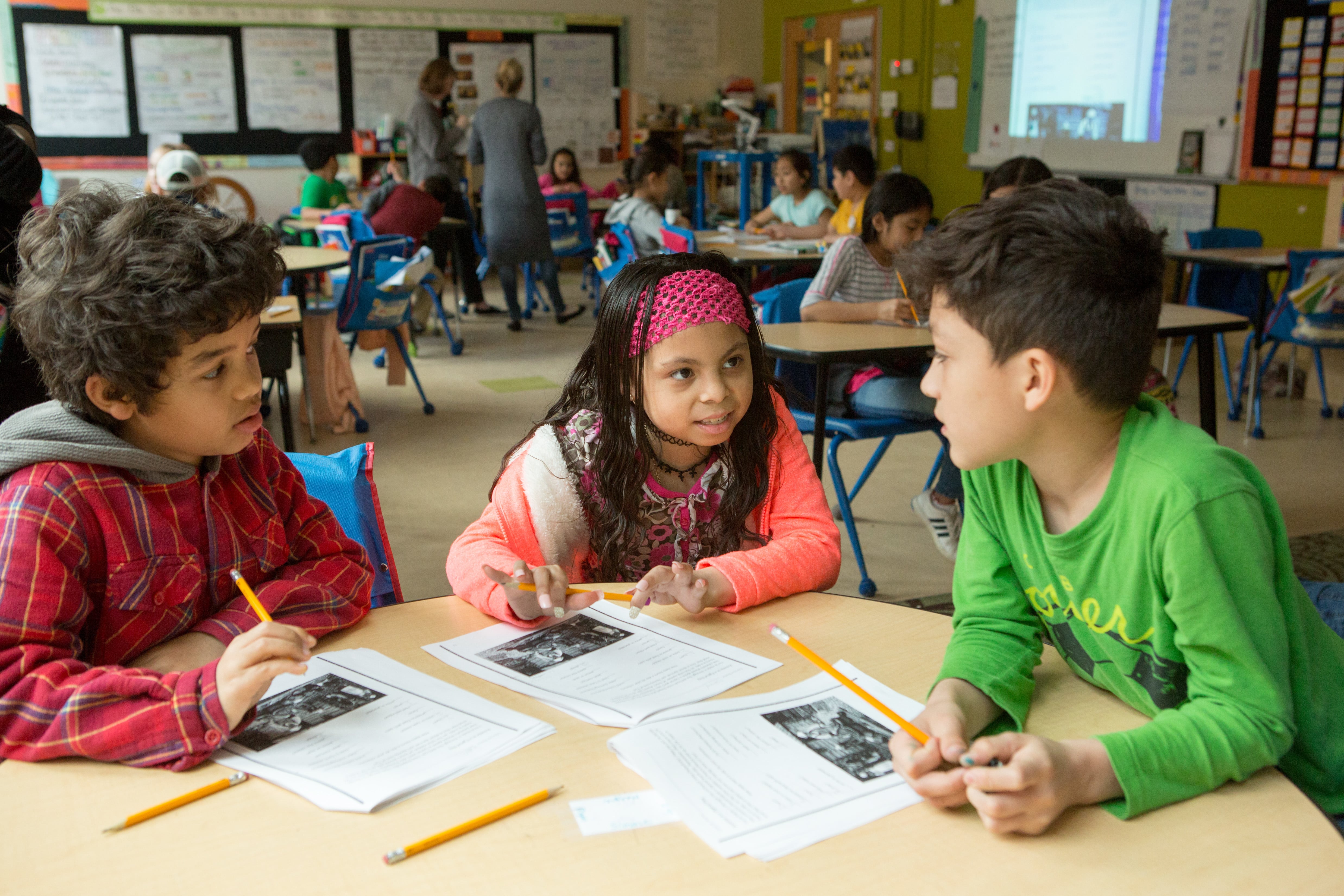 Two young boys and a girl sit at a table in a classroom with pencils and worksheets.