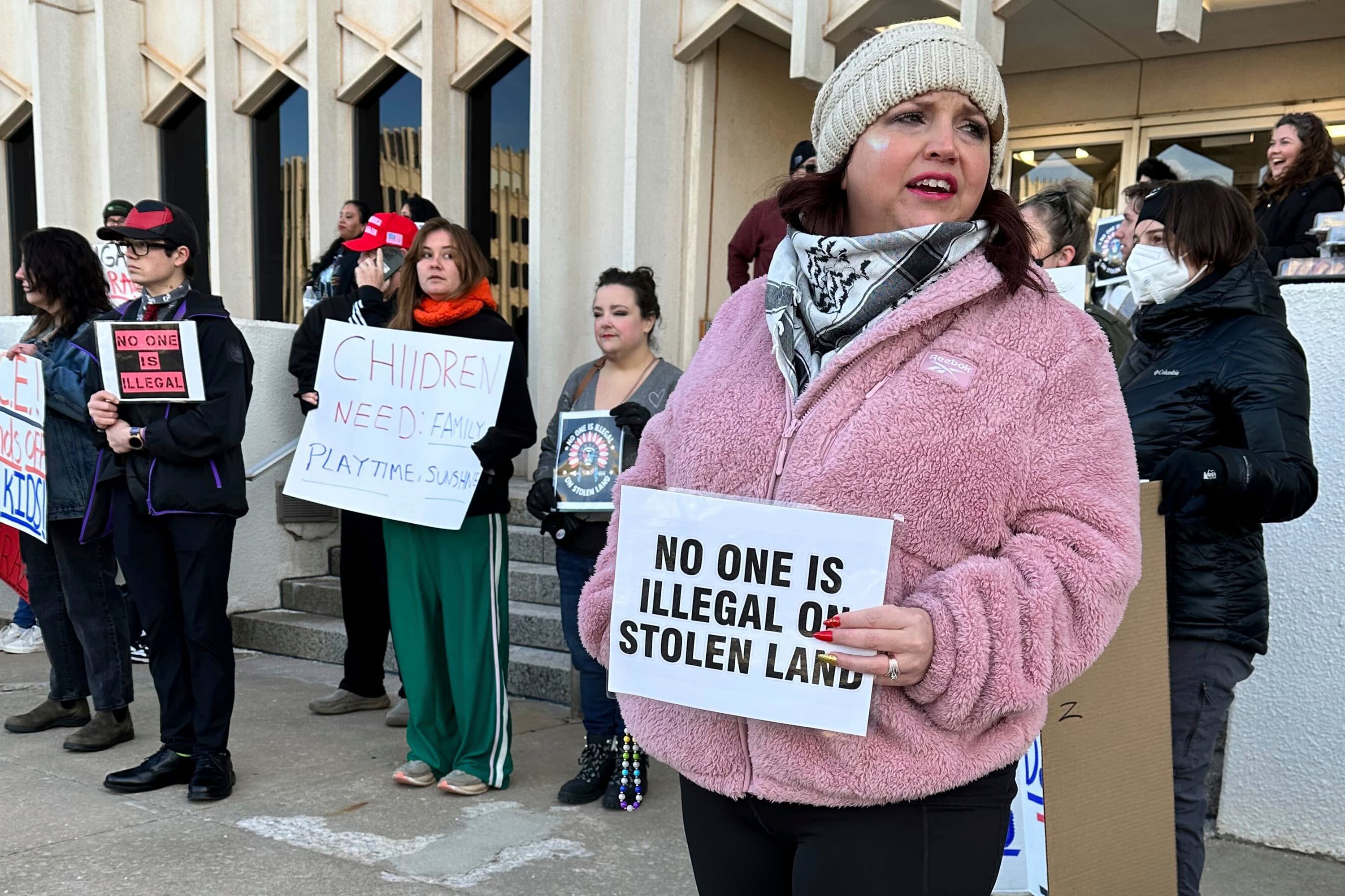 A woman in a pink sweater and holding a sign stands in front of a large building and a group of people holding signs and protesting outside.
