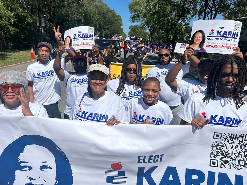 A group of people wearing white campaign t-shirts and holding signs stand outside.
