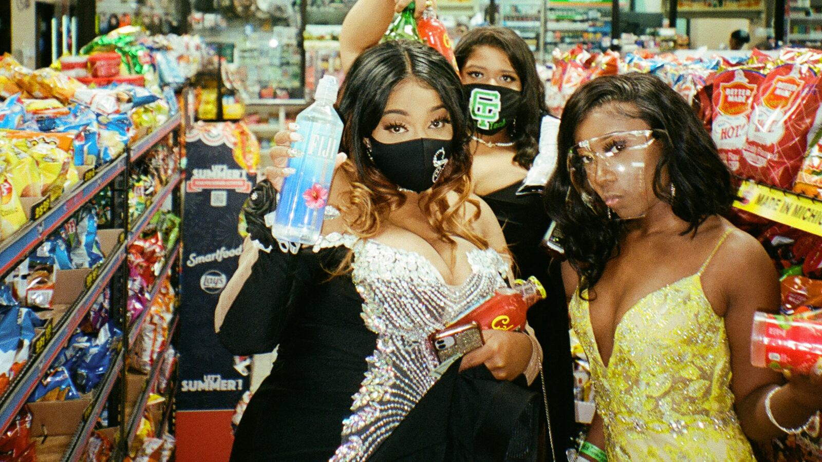 Three young women pose for a picture together wearing their ornate prom dresses and protective masks in a convenience store.