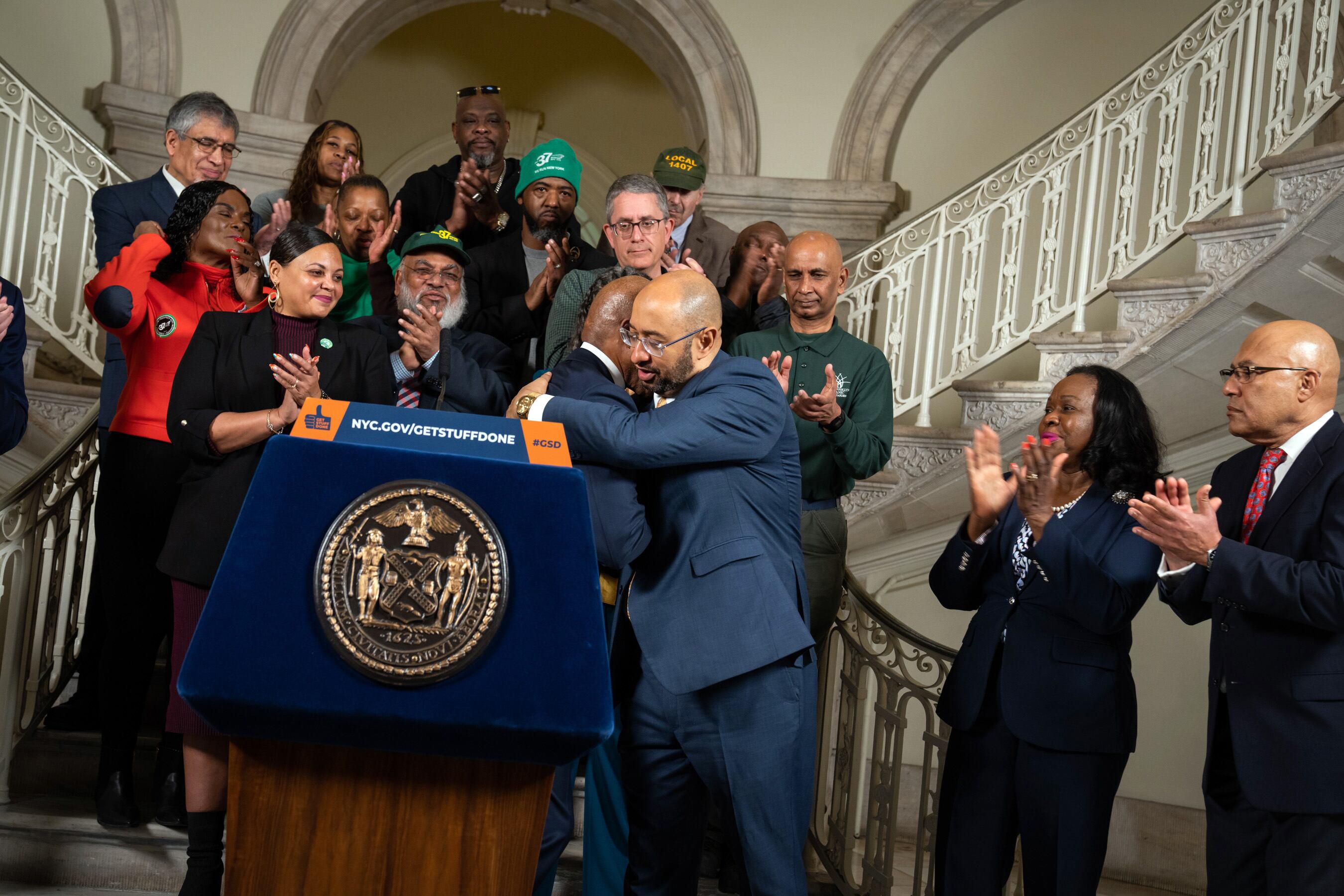 Two men in suits embrace next to a podium surrounded by people.