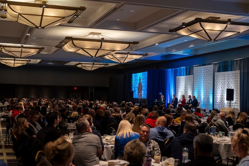 A photograph of a large audience watching five adults wearing suits sitting on stage.