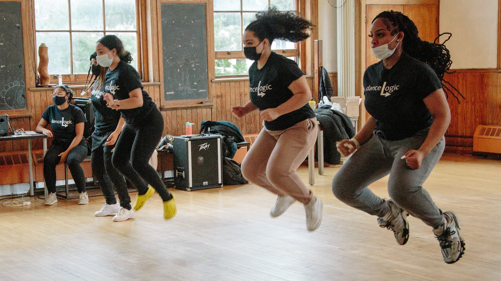 Girls wearing black shirts and face masks jump high over a wooden floor during class.