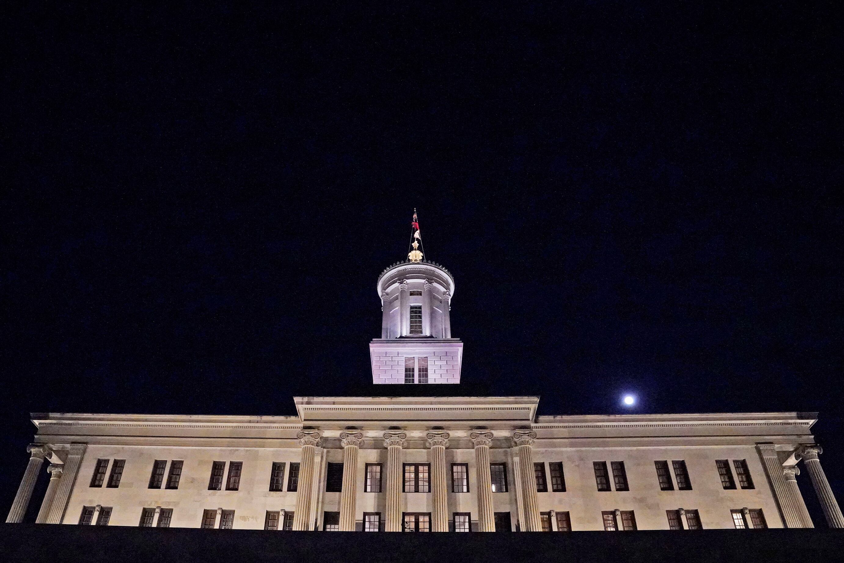 An illuminated tan stone building with a flag at the top with a dark night sky in the background.