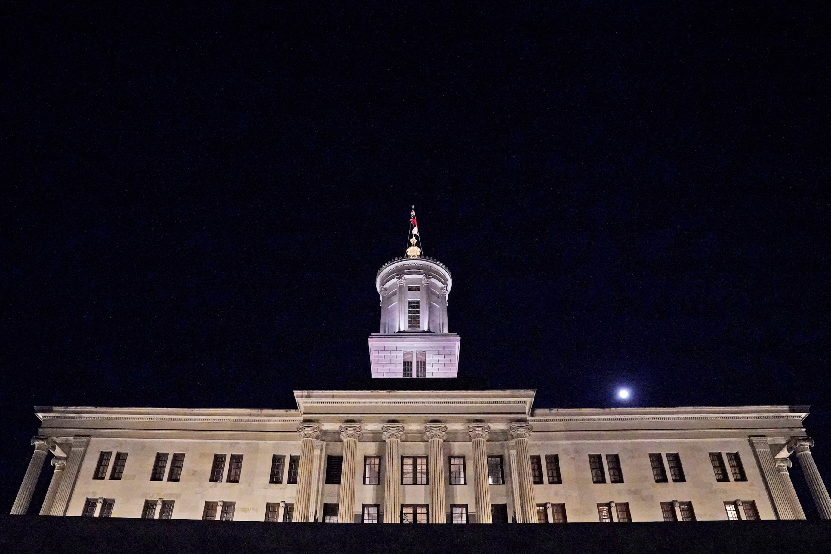 An illuminated tan stone building with a flag at the top with a dark night sky in the background.