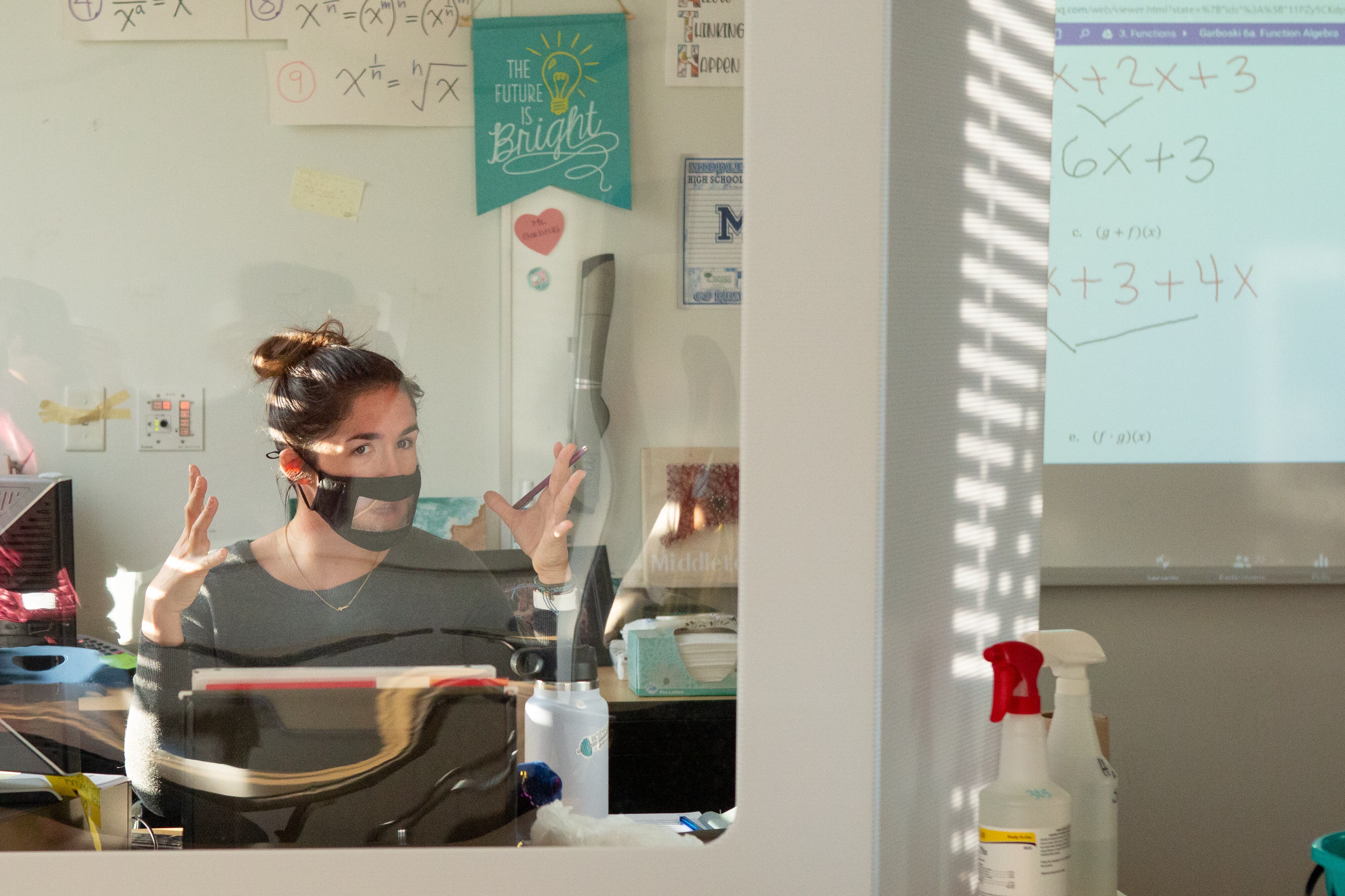 A math teacher wearing a mask sits behind her desk, which is protected by a large glass shield due to COVID-19 procedures.