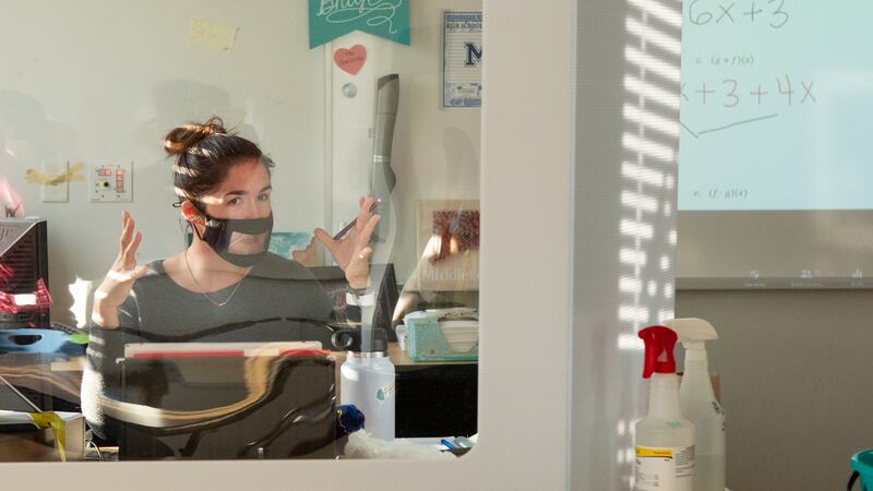 A math teacher wearing a mask sits behind her desk, which is protected by a large glass shield due to COVID-19 procedures.