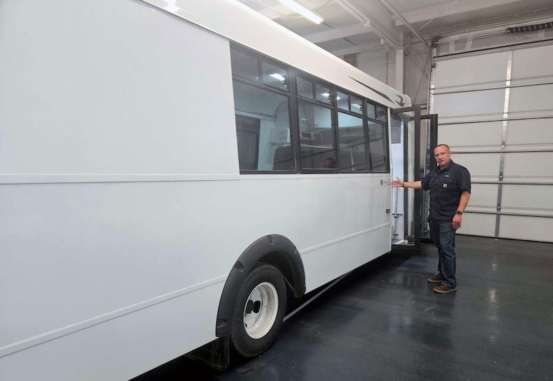A person wearing a dark blue shirt and jeans stands at the entrance of an all-white bus.