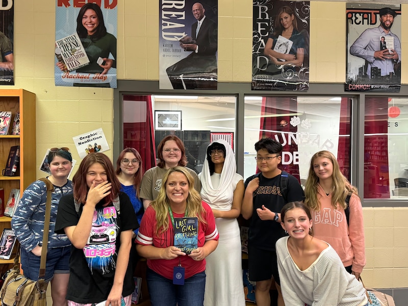 A teacher in a red shirt stands with a group of students in the school library, holding a book called Last Girl Breathing.