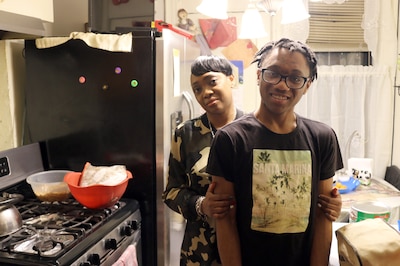 A Black mother and her son pose for portraits side by side in their home. The teenaged son is wearing glasses and the mom is wearing camouflage sweater. They are standing in their kitchen next to a stove and fridge.