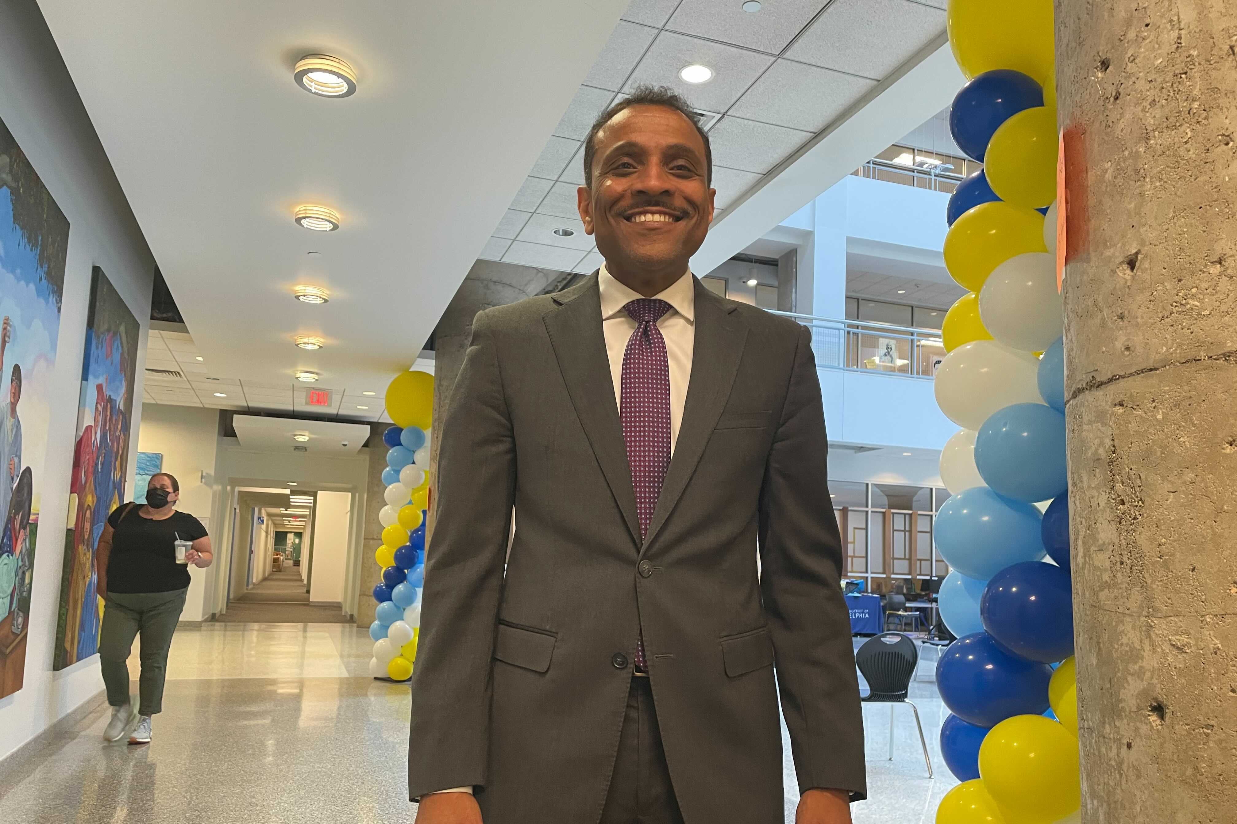 A smiling man in a gray suit stands in a hallway as a woman walks behind him. There are blue, yellow, and white balloons to his left.