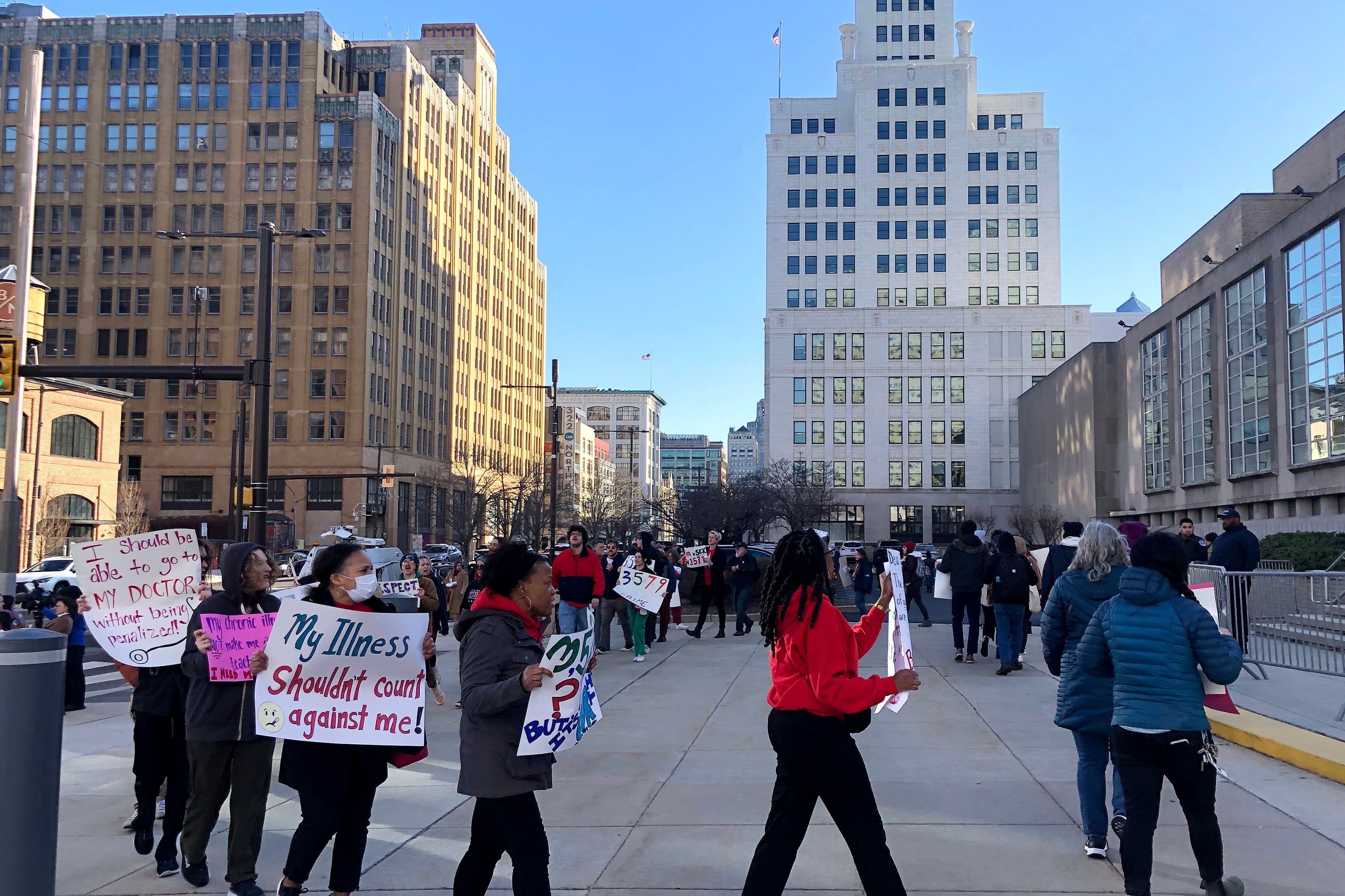 A large group of people holding signs march in a circle outside the front entrance to a large building with tan stone and many windows.