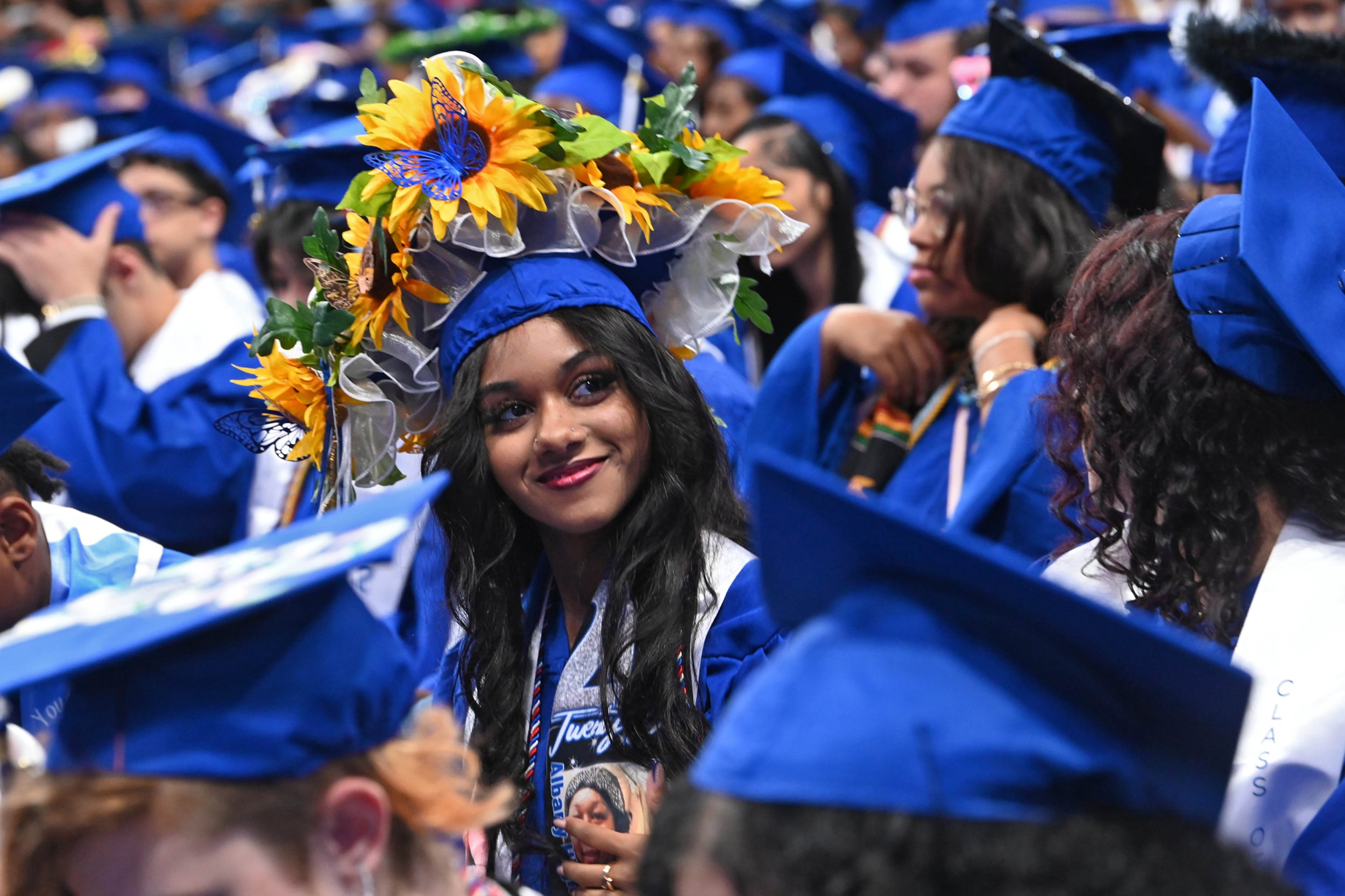A young woman with long dark hair and wearing flowers out of her blue graduation cap is surrounded by a sea of high school graduates all in blue graduations caps and gowns.