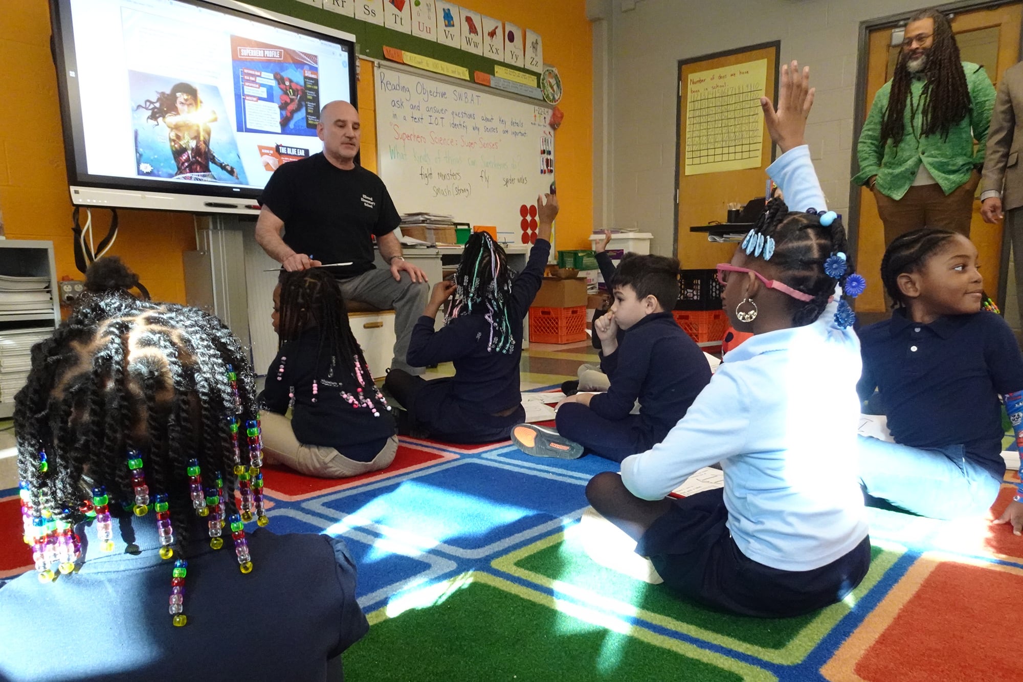 Adults stand near a group of young students while some have their hands raised and sitting on a colorful rug in a classroom.