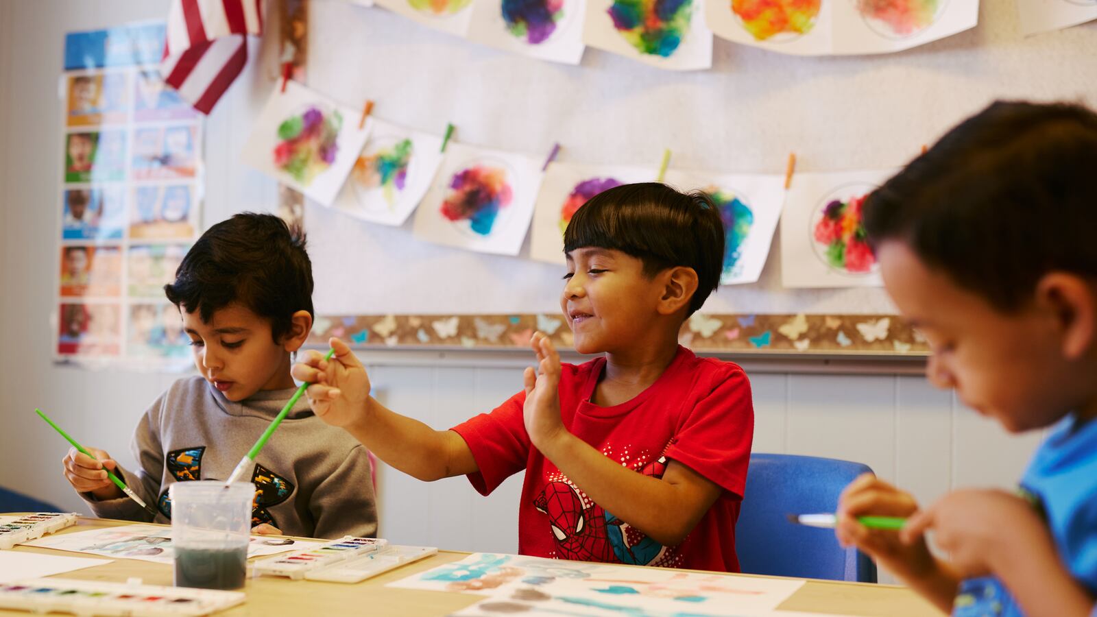 Three young students paint during preschool class on a wooden table with art hanging to dry in the background.