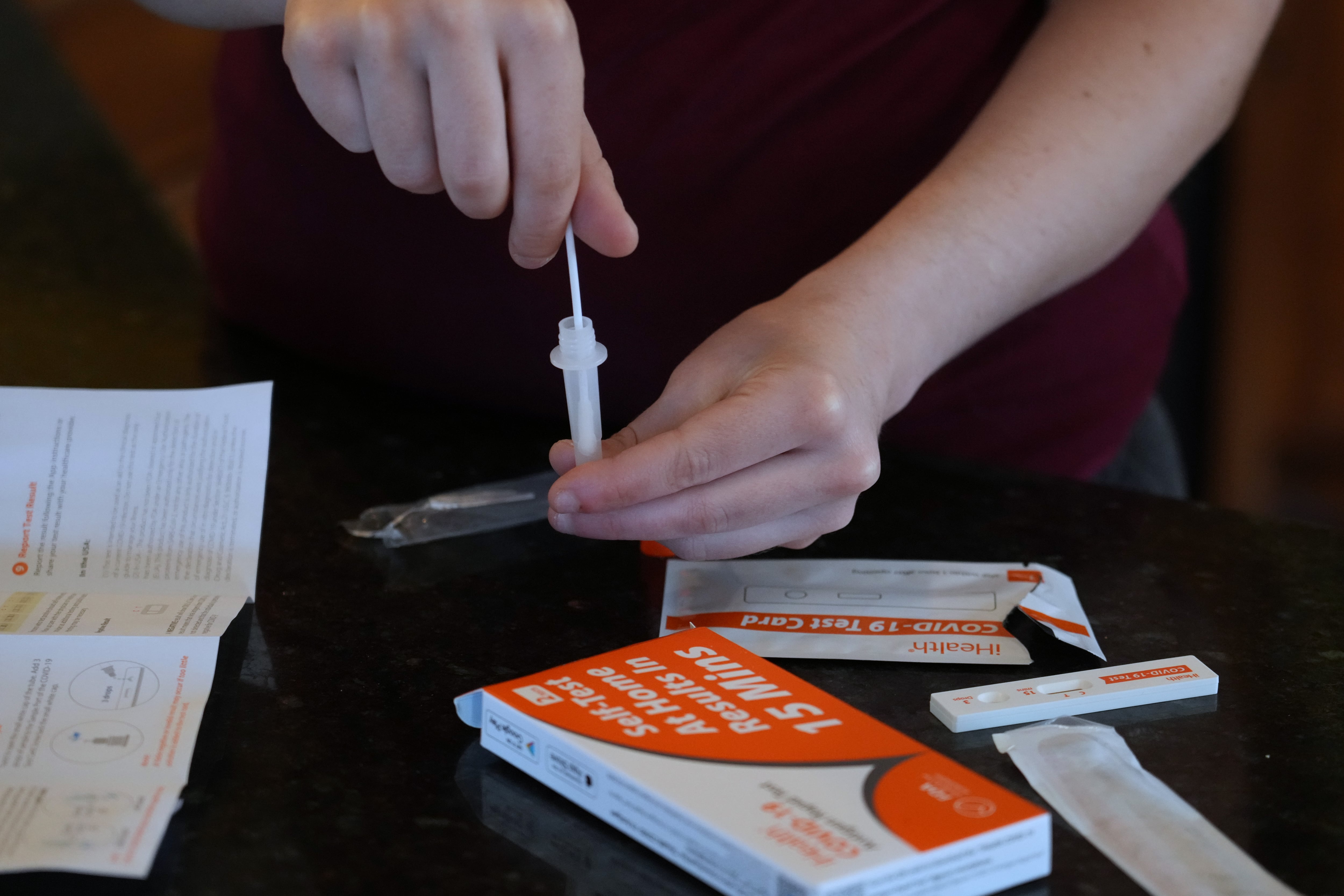 A COVID-19 at-home rapid test package is opened on a table as someone dips a swab into the tube that is included with the kit.