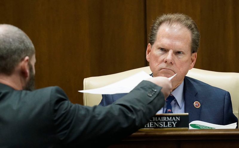 Inside of a paneled legislative chamber, a man wearing a suit looks somber as he receives paperwork from another man.