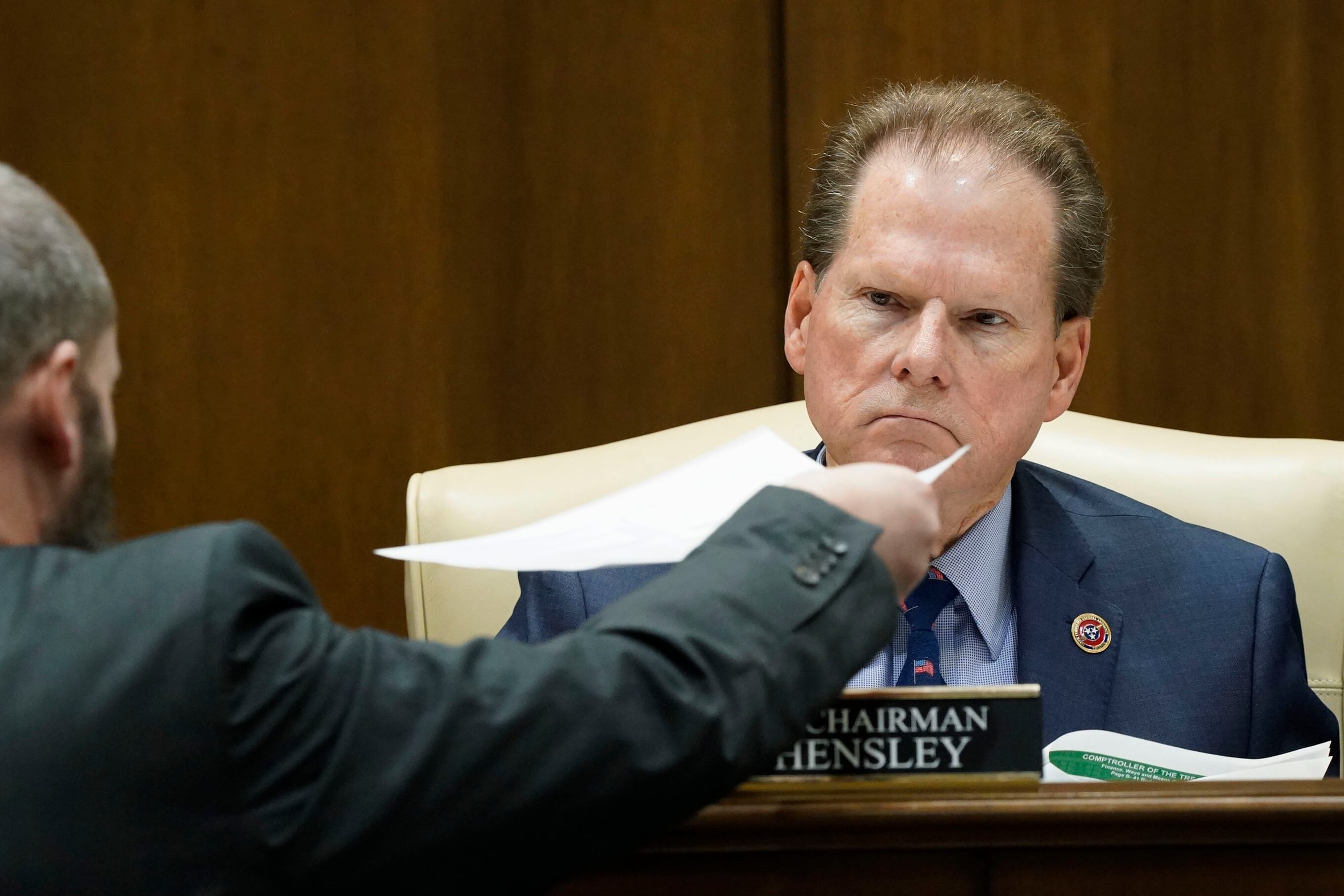 Inside of a paneled legislative chamber, a man wearing a suit looks somber as he receives paperwork from another man.