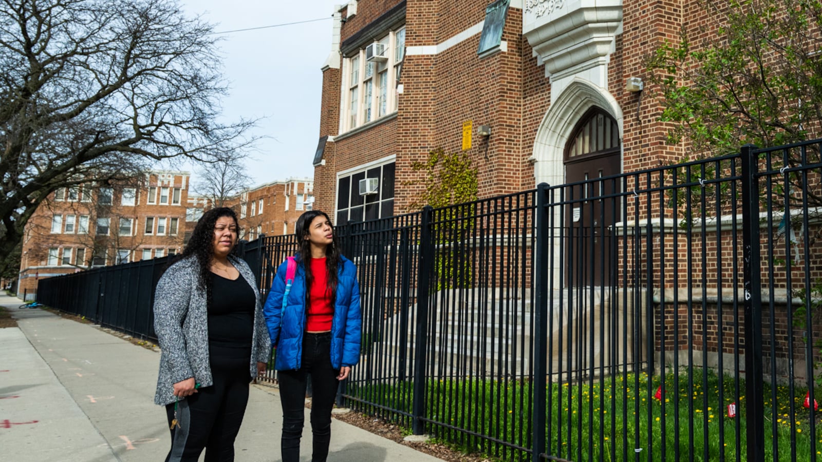 An adult woman and her daughter, both with dark hair and wearing jackets, stand outside of a fenced in brick school building.