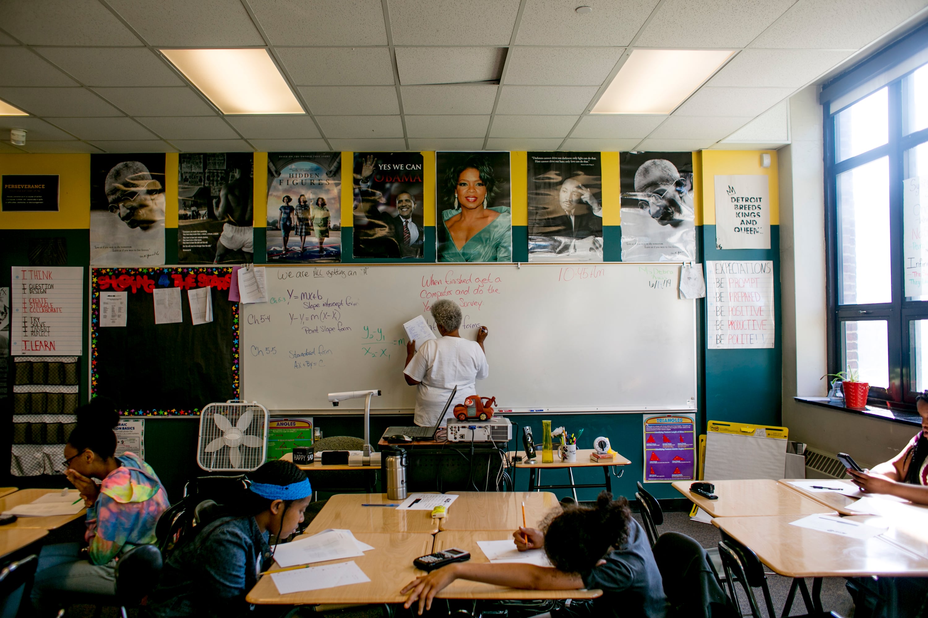 A teacher writes on the board with students at their desks in the foreground.